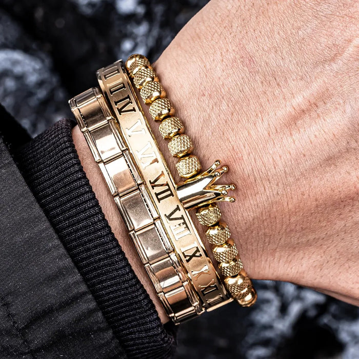 A gold-colored three-piece bracelet set on a man's wrist, with one bracelet featuring a crown decoration.